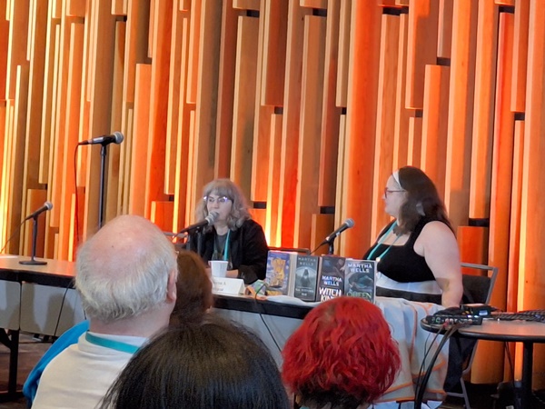 Slightly blurry photo of a table at the front of a room with vertical orange wooden design behind it. At the table sits Martha Wells, speaking into the microphone, and the interviewer (whose name I forgot) sitting beside her.